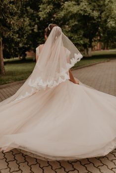 dark-haired European Caucasian young woman bride in white wedding dress with long veil and tiara on head spinning whirls dancing posing moving at nature on background greenery. Valentine love couple