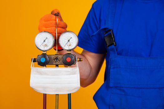 An air conditioning technician holds pressure gauges in his hand for refueling air conditioners. Medical face shield on measuring equipment for filling air conditioners.