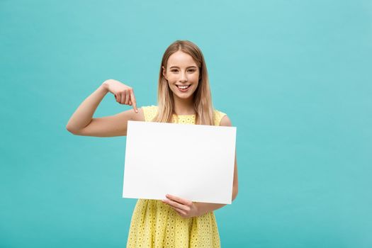Portrait of young woman in yellow dress pointing finger at side white blank board. Isolated over Blue background