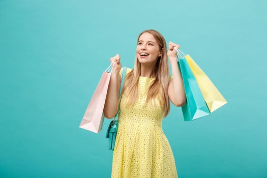 Lifestyle Concept: Portrait of shocked young attractive woman in yellow summer dressposing with shopping bags and looking at camera over blue background
