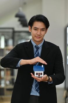 Portrait of asian man real estate agent hand holding house model and smiling to camera. New house, insurance and real estate concept.