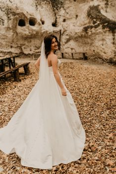 young woman bride in white wedding dress with long veil run away through autumn forest on fallen orange leaves to mountain. brunette with short hair bob