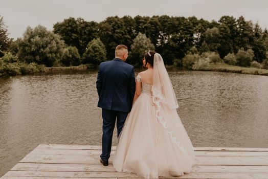 blonde European Caucasian young man groom in blue suit and black-haired woman bride in white wedding dress with long veil and tiara on head. Newlyweds kissing hugging on wooden pier bridge on lake