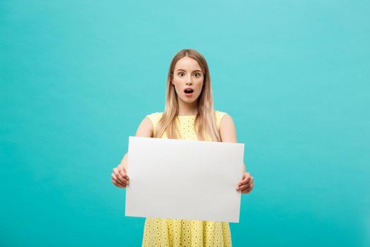 Portrait of amazed young blond woman holding blank sign with copy space on blue studio background. Showing shocked surprise face