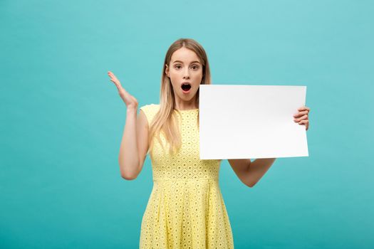 Closeup portrait, young woman in yellow dress holding white plain paper with shocked expression at what she sees, isolated blue background