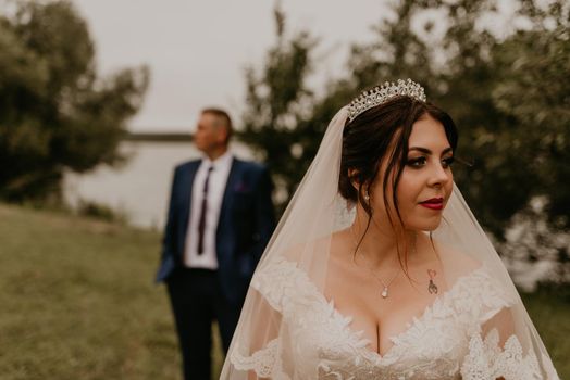 European Caucasian young man groom in blue suit and black-haired woman bride in white wedding dress with long veil and tiara on head. newlyweds look in different directions. man stands behind woman