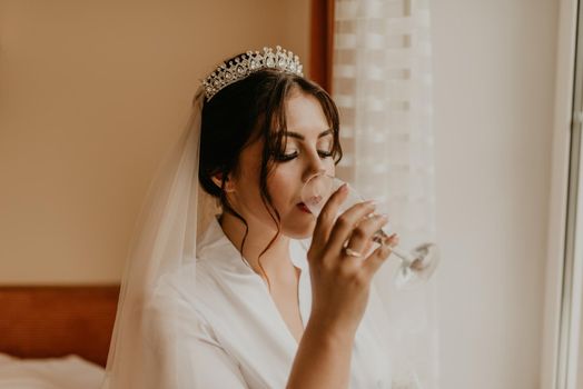European Caucasian young black-haired woman bride white silk peignoir thin robe long veil and tiara on head stand near window . morning gatherings of drinking champagne preparing bride ceremony