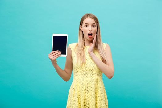 Portrait of a surprised amazed young woman looking at digital tablet isolated on a blue studio background