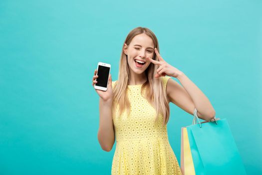 Portrait young attractive woman with shopping bags shows the phone's screen directly to the camera. Isolated on blue background.