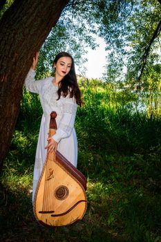 Ukrainian woman in a white dress stands under a tree by the river. Ukrainian musician with a national stringed bandura.