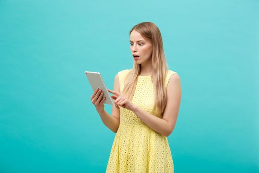 Portrait of a surprised amazed young woman looking at digital tablet isolated on a blue studio background