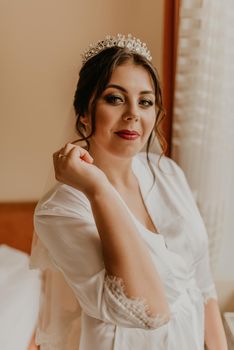 European Caucasian young black-haired woman bride white silk peignoir thin robe long veil tiara on head. morning gatherings of preparing bride for holiday party wedding. girl sitting on bed in bedroom