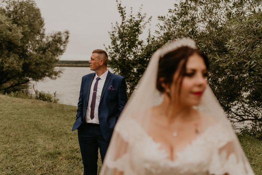 European Caucasian young man groom in blue suit and black-haired woman bride in white wedding dress with long veil and tiara on head. newlyweds look in different directions. man stands behind woman