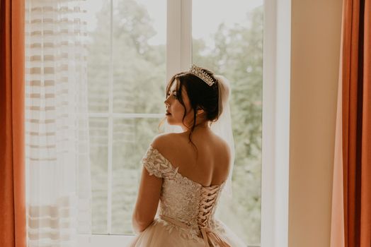 European Caucasian young black-haired woman bride in white wedding dress with long veil and tiara on head. girl stands with her back near window morning gatherings of preparing bride for holiday party