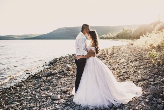 groom and bride in wedding dress stand in embrace on rocky bank near river in summer outdoors In rays of setting sun. Bakota, Ukraine