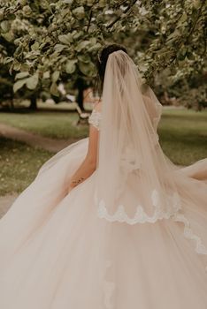 dark-haired European Caucasian young woman bride in white wedding dress with long veil and tiara on head spinning whirls dancing posing moving at nature on background greenery. Valentine love couple