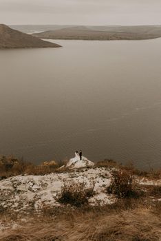 a wedding couple in love, groom and the bride in dress veil stand on Big Stone on cliff and kiss. panoramic view big river lake sea in distance silhouettes of islands. bakota ukraine europe