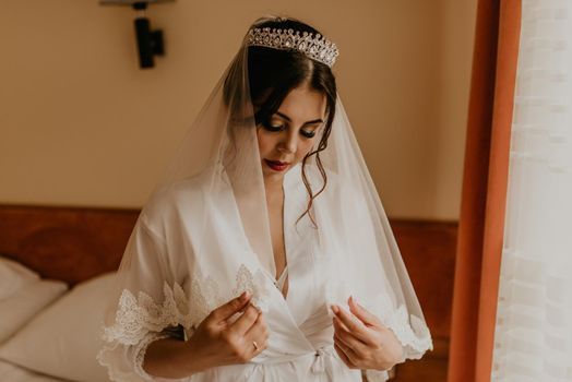 European Caucasian young black-haired woman bride white silk peignoir thin robe long veil tiara on head. morning gatherings of preparing bride for holiday party wedding. girl sitting on bed in bedroom