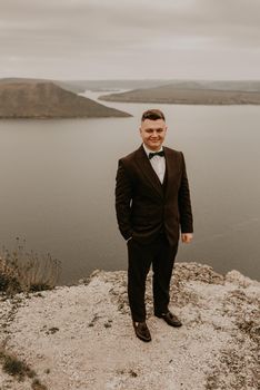 adult man with short hair in classic suit, white shirt, bow tie on neck. lonely person in nature outdoor. panoramic view big river lake sea in distance silhouettes of islands. bakota ukraine