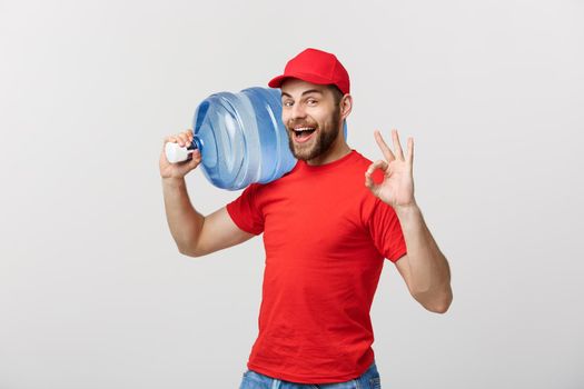 Portrait smiling bottled water delivery courier in red t-shirt and cap carrying tank of fresh drink and showing ok isolated over white background