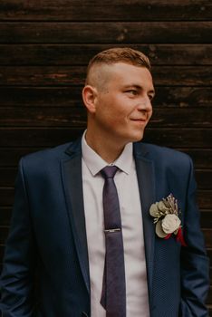 blonde European Caucasian young man groom in navy blue suit white shirt and tie looking to the side. flower boutonniere attached to a breast pocket