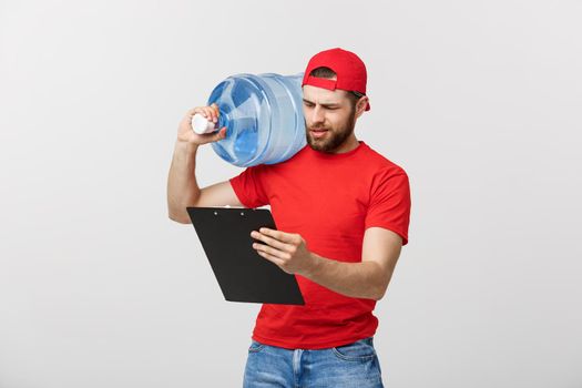 Portrait smiling bottled water delivery courier in red t-shirt and cap carrying tank of fresh drink and showing thumb up isolated over white background.