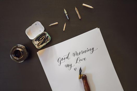 Good morning. Calligrapher Young Woman writes phrase on white paper. Inscribing ornamental decorated letters. Calligraphy, graphic design, lettering, handwriting, creation concept.