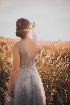 The young beautiful girl in a gray dress with an open back raised her hands up and closes the rays of the sun. against the background of blurred tall yellow grass at sunset.