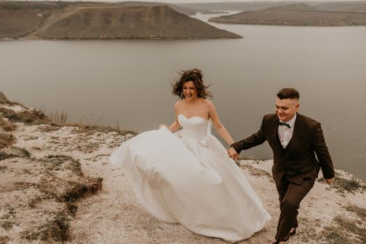 loving couple wedding newlyweds outdoor. bride in white dress long veil and groom in suit walk in summer fall on mountain above river. sunrise. man and woman on rocks above cliff. bakota ukraine