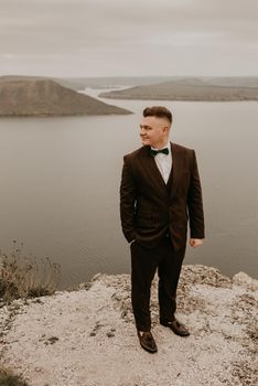 adult man with short hair in classic suit, white shirt, bow tie on neck. lonely person in nature outdoor. panoramic view big river lake sea in distance silhouettes of islands. bakota ukraine