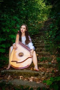 A Ukrainian woman is sitting on the stairs in an old park with a Ukrainian bandura musical instrument.