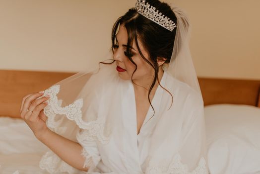 European Caucasian young black-haired woman bride white silk peignoir thin robe long veil tiara on head. morning gatherings of preparing bride for holiday party wedding. girl sitting on bed in bedroom