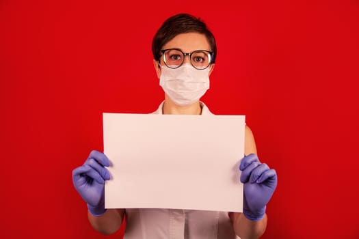 A nurse in a medical protective mask holds a blank white sheet on a red background. The concept of protection against viruses, influenza and coronavirus COVID-19. Health care and surgical concept.