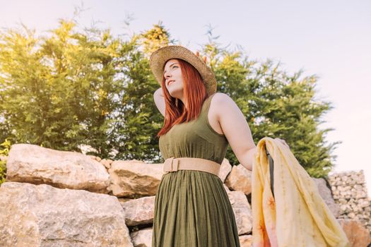 adventurous young girl, relaxing on her summer holiday. woman on a trip to the beach strolling along the coast. bathed in sunlight. woman in green summer dress, straw hat, outdoors with natural light and plants in background.