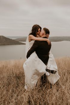 loving couple wedding newlyweds in white dress veil sports shoes and suit hug kissing whirl on tall grass in summer field on mountain above the river. sunset.