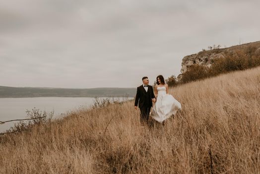 A loving couple wedding newlyweds in a white dress and a suit walk hug kissing on the tall grass in the summer autumn field on the mountain above the river. sunset. bakota ukraine