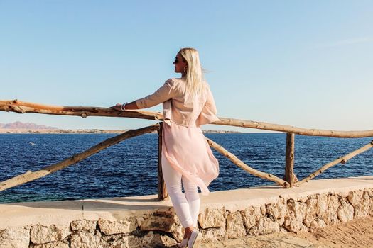 Beautiful woman blonde stands near the railing of the pier and looks at the sea. Rear view of a young woman looking at the sea in a resort
