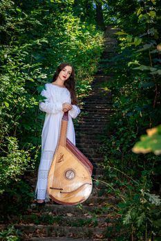 Ukrainian girl in a white dress stands on a stone ladder in the middle of the forest. Ukrainian woman with loose braids with a bandura musical instrument.