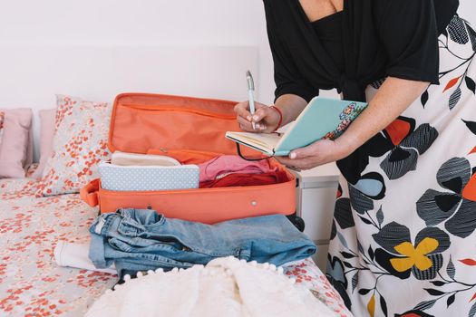 cropped shot of a mature blonde woman, jotting down in her diary the items of luggage she is taking for her trip. business woman preparing her trip. bedroom room, natural light, clothes and travel items on the bed.