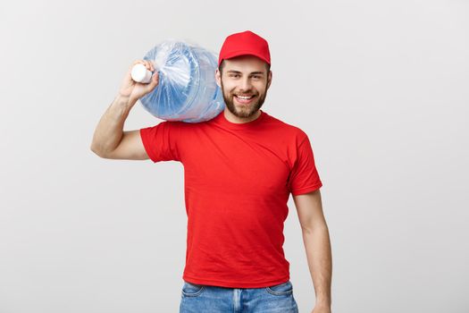 Delivery Concept: Portrait of smiling bottled water delivery courier in red t-shirt and cap carrying tank of fresh drink isolated over grey background.