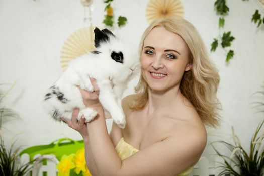 A lovely young blond woman standing in profile closeup and holding a young bunny in her arms. Beautiful blonde girl holding a white rabbit in her arms. Easter bunny sitting in the arms of a girl