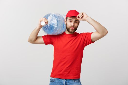 Delivery Concept: Portrait of smiling bottled water delivery courier in red t-shirt and cap carrying tank of fresh drink isolated over grey background.