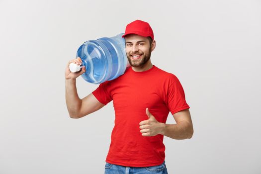Portrait smiling bottled water delivery courier in red t-shirt and cap carrying tank of fresh drink and showing thumb up isolated over white background.