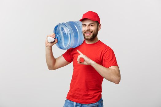 Portrait smiling bottled water delivery courier in red t-shirt and cap carrying tank of fresh drink and pointing finger isolated over white background