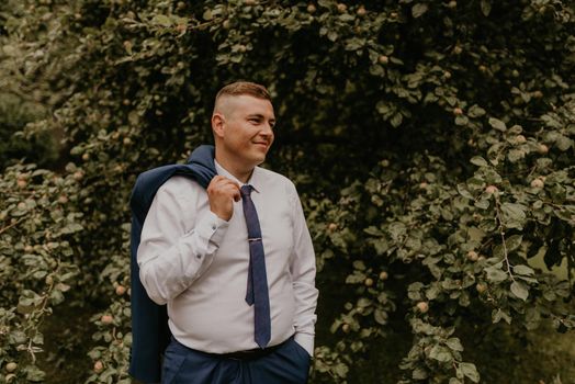 young blond man European curly-haired successful businessman groom in classic clothes white shirt and tie stands on street holding jacket thrown over his back shoulder in nature green leafy wall