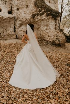 young woman bride in white wedding dress with long veil run away through autumn forest on fallen orange leaves to mountain. brunette with short hair bob