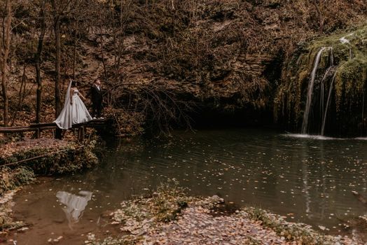 bride in dress and long veil groom in suit stand on wooden pier above water. small pond lake river. summer-autumn sunset. waterfall Burbun in village Lysets Dunaivtsi Khmelnytskyi