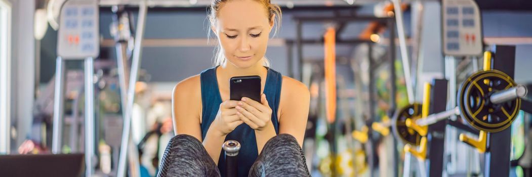 Young woman using phone while training at the gym. Woman sitting on exercising machine holding mobile phone. BANNER, LONG FORMAT
