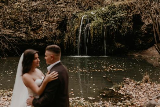 couple out of focus. skinny bride in white wedding dress and long veil hugs groom in a suit. small pond lake river. summer-autumn sunset fallen orange leaves mist. waterfall Burbun in village Lysets Dunaivtsi Khmelnytskyi