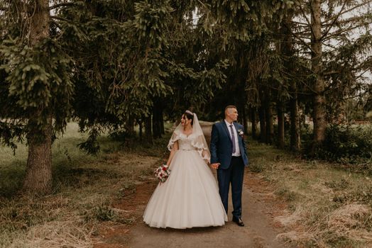 blonde European Caucasian young man groom in blue suit and black-haired woman bride in white wedding dress with long veil and tiara on head. newlyweds look in different directions holding hands